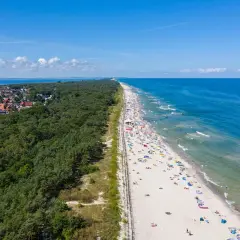 Breiter Sandstrand mit vielen Badegästen, gesäumt von einem dichten Kiefernwald und einer kleinen Stadt. Ein echter Geheimtipp an der polnischen Ostsee.