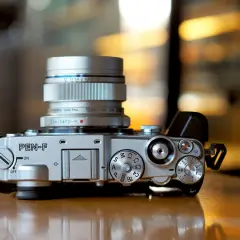 Silver Olympus PEN-F camera with a silver Olympus Digital 12mm 1:2.0 lens attached. The camera body features various dials and buttons, ready for capturing stunning images with its Olympus PEN F lenses.