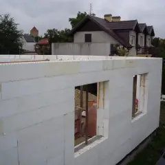 Budowa domu ze styropianu. Białe bloki tworzą ściany z otworami okiennymi. W tle widać inne budynki i drzewa.