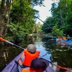 Rodzina płynie kajakiem po malowniczym Pojezierzu Brodnickim, otoczona bujną zielenią.