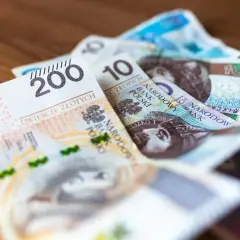 Stos banknotów, w tym 200 i 10 złotych, leży na drewnianym blacie. To skróty na pasku wypłaty.
