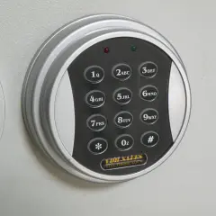 Elektroniczna klawiatura szyfrowa sejfu z przyciskami cyfrowymi i symbolami. W tle widoczne są metalowe tuleje mocujące.