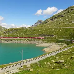 Ett rött tåg slingrar sig längs en turkos sjö i en grönskande bergsmiljö. En resa med tåg i Italien kan se ut så här.