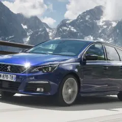 Niebieski Peugeot 308 T9 kombi jedzie po górskiej drodze, z ośnieżonymi szczytami w tle.