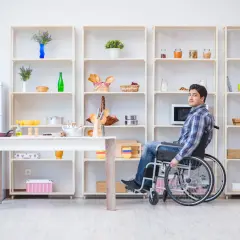 Mężczyzna na wózku inwalidzkim w kuchni, która jest przystosowana jako mieszkanie dla niepełnosprawnych. Wygodny dostęp do półek i stołu.