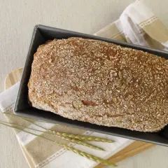 Świeżo upieczony chleb orkiszowy w keksówce, posypany ziarnami. Obok leżą kłosy zbóż i lniana ściereczka.