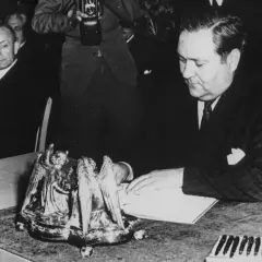 Carlo Schmid sitzt an einem Tisch und blickt auf ein Buch. Ein Fotograf hält eine Kamera.
