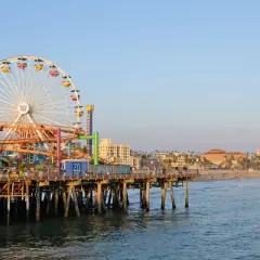Der Santa Monica Pier mit seinem Riesenrad und Achterbahn ragt ins Meer. Im Hintergrund erstreckt sich der sonnige los angeles strand mit Palmen und Gebäuden.