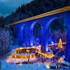 Magischer weihnachtsmarkt deutschland geheimtipp unter beleuchteter Brücke. Ein Zug fährt vorbei, während Besucher Buden und einen Weihnachtsbaum bestaunen.