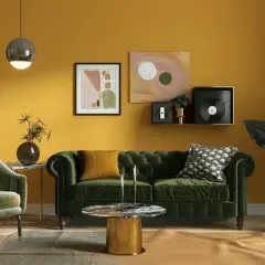 Ein Wohnzimmer mit senfgelben Wänden. Ein grünes Samtsofa und ein Sessel. Welcher Farbe passt zu Gold? Der goldene Couchtisch und Akzente im Bild zeigen, dass Grün und Gelb gut harmonieren.