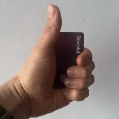 Dłoń trzymająca fioletową kartę Revolut, kciuk uniesiony w geście aprobaty.