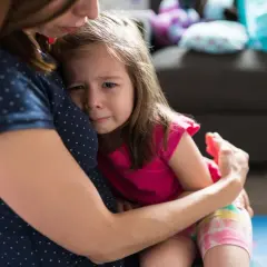 Mała dziewczynka z zapłakaną buzią mocno przytula się do mamy, wyrażając swój lęk separacyjny.