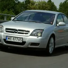 Srebrny Opel Vectra jedzie po drodze, za nim zielone drzewa.