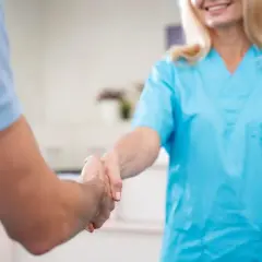 Uśmiechnięta opiekunka w niebieskim fartuchu wita pacjenta uściskiem dłoni. Kiedy należy się opiekunka z mops?