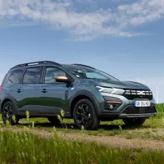 Un'auto familiare verde scuro, con barre sul tetto e dettagli color rame, parcheggiata su un sentiero sterrato in un campo con mulini a vento sullo sfondo.