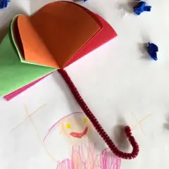 Buntes Papier und Pfeifenreiniger ergeben einen tollen Regenschirm zum Basteln. Kleine blaue Tupfer symbolisieren Regentropfen.