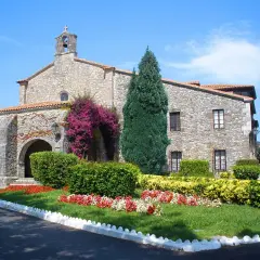 El **santuario de la Barquera**, con su fachada de piedra y arcos, se adorna con vibrantes buganvillas y un jardín florido bajo un cielo azul.