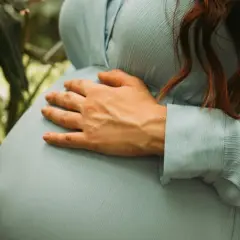 Gravid i vecka 5, en kvinna med ljusblå klänning håller om sin mage bland gröna blad.