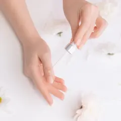 Dłonie z pipetką aplikującą olejek na paznokieć, otoczone białymi kwiatami.