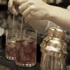 Barkeeper mixt einen roten Bier Cocktail mit Eis in einem Glas. Daneben steht ein goldener Shaker.