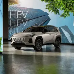 Srebrny SUV RAV4 HEV stoi przed ekranem z nowoczesnymi budynkami i zielenią.