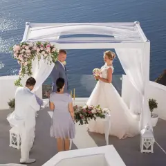 Piękny ślub w Grecji. Para młoda stoi pod białym baldachimem, z widokiem na błękitne morze. W tle widać skaliste wybrzeże.
