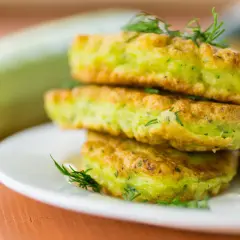 Frittelle di zucchine dorate e soffici, guarnite con aneto fresco, impilate su un piatto bianco. Sullo sfondo una zucchina intera.
