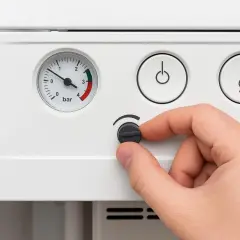 Dłoń reguluje pokrętłem piec gazowy. Manometr wskazuje niski poziom ciśnienia, sugerując brak wody w instalacji.