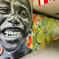 Graffiti-Porträt eines lachenden Kindes neben einer bunten Wand mit Schriftzügen.