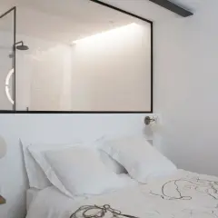 Stanza buia con letto bianco, una finestra tra due stanze rivela un bagno moderno con doccia e specchi luminosi.