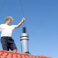 Pracownik na dachu czyści komin domu za pomocą szczotki.