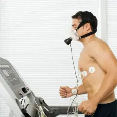 Mężczyzna w masce biegający na bieżni, podłączony do aparatury EKG. Wyniki testu VO2max w tabeli na ekranie bieżni.