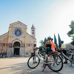 A group enjoys a sunny Verona bike tour, with bicycles parked in front of a grand cathedral and a medieval tower.