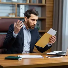 Homme surpris ouvrant une lettre de contestation de facture, assis à son bureau avec un ordinateur portable.