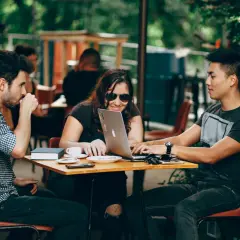 Tres jóvenes colaboran en un portátil en una cafetería al aire libre, un ejemplo de cómo ser nómada digital.