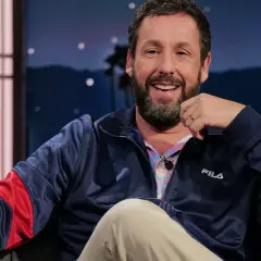 Adam Sandler skrattar glatt i en blå Fila-jacka, redo för en rolig intervju.