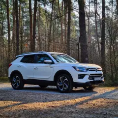 Biały SsangYong Korando zaparkowany na leśnej drodze, otoczony przez wysokie sosny.