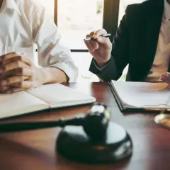 Mężczyzna w białej koszuli, pozwany, siedzi z rękami złożonymi na otwartej księdze. Obok młotek sędziowski i waga sprawiedliwości.