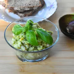 Pyszna pasta jajeczna z awokado, udekorowana bazylią, obok połówka awokado i kromki chleba.