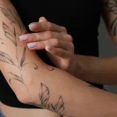 Delikatny tatuaż gałązki z liśćmi na przedramieniu. Dłonie z jasnym manicurem dotykają skóry, podkreślając, ile goi się tatuaż.
