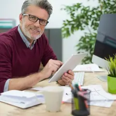 Uśmiechnięty mężczyzna w okularach pracuje na tablecie, przeglądając dokumenty. Może to być jego nowy kontrakt menedżerski.