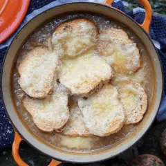 Zuppa di cipolle fumante in una casseruola arancione, con crostini dorati e formaggio fuso. Accanto, pane tostato e cipolle crude.