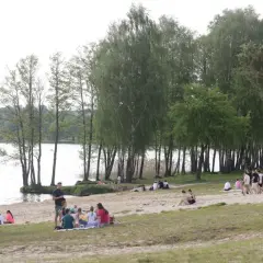 Ludzie odpoczywają na piaszczystej plaży nad jeziorem Pławniowice, ciesząc się wiosennym dniem wśród drzew.