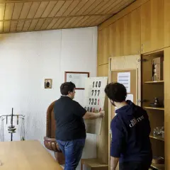 Zwei Männer im katholischen Priesterseminar hängen eine Liste mit Zahlen auf. Ein Kruzifix hängt an der Wand.