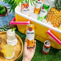 Letnia piknikowa atmosfera z drinkami Bacardi. W żółtej lodówce schłodzone puszki rumowych koktajli i ananas. Na trawie dwa drinki, kokosy i limonki.