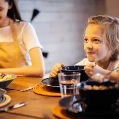 Uśmiechnięty chłopiec je z miseczki, podczas gdy mama przygotowuje kanapkę. Rodzina cieszy się wspólnym, dobre jedzenie przy drewnianym stole.