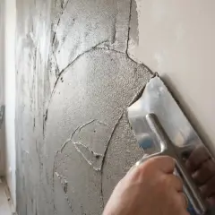 Ręka z kielnią rozprowadza masę naprawczą na ścianie. W tle okno zabezpieczone folią.