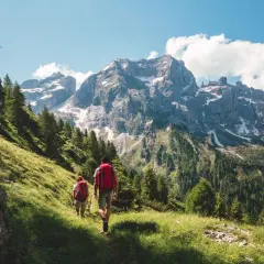 Dos personas disfrutan del senderismo y trekking por un sendero verde con imponentes montañas nevadas de fondo.