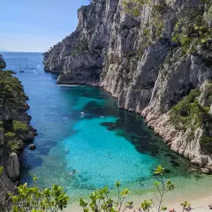 Aguas turquesas y cristalinas en el **calanque d'en Vau**, rodeado de imponentes acantilados rocosos y vegetación. Gente nadando y barcos a lo lejos.