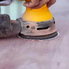 Mano che usa una levigatrice orbitale per carteggiare il legno verniciato, preparando la superficie per una nuova finitura.